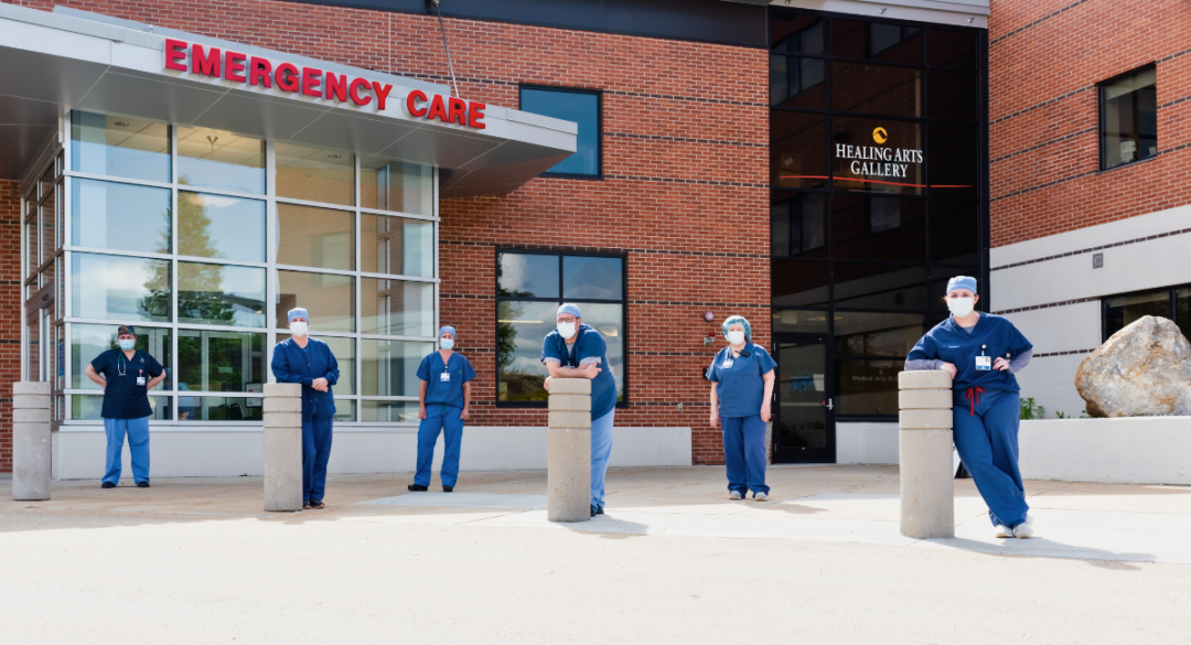 Six healthcare professionals in scrubs and masks stand in front of the Emergency Care entrance of a hospital, with a brick building and large glass windows in the background. A sign for the Healing Arts Gallery is visible on the building