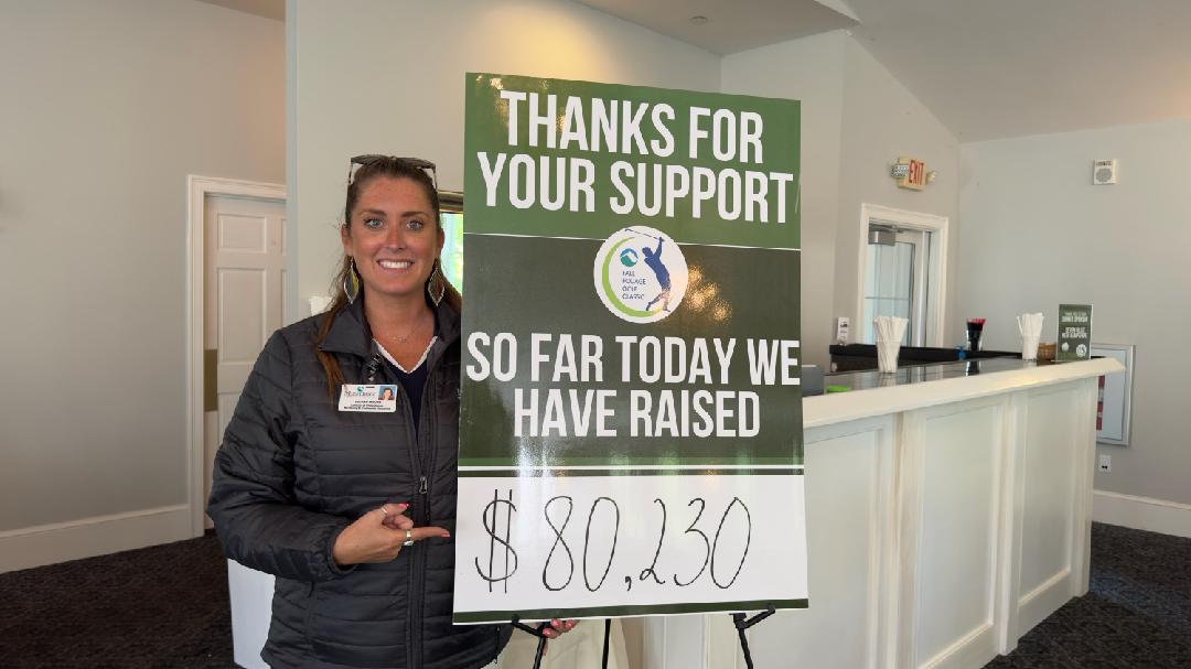 LeeAnn Clark Moore stands next to a sign that reads, "Thanks for your support. So far today we have raised $80,230." She is smiling and pointing at the amount raised on the sign.