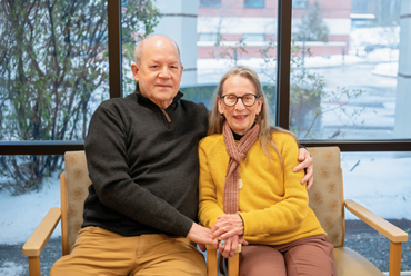 Craig and Judy Unger-Clark sit together smiling in a hospital waiting area with snow outside the window. Craig wears a dark sweater; Judy is in a yellow sweater and scarf.