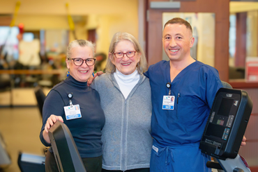 Rosamond Cady stands between two smiling healthcare providers—one woman in glasses and a black turtleneck with a name badge, and one man in blue scrubs with an MD badge. They are in a bright, welcoming rehabilitation or wellness area with fitness equipment visible in the background.