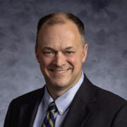 Professional portrait of a man with short hair, wearing a dark suit and striped tie, smiling against a gray textured background