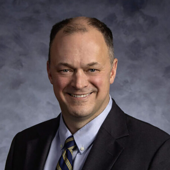 Professional portrait of a man with short hair, wearing a dark suit and striped tie, smiling against a gray textured background