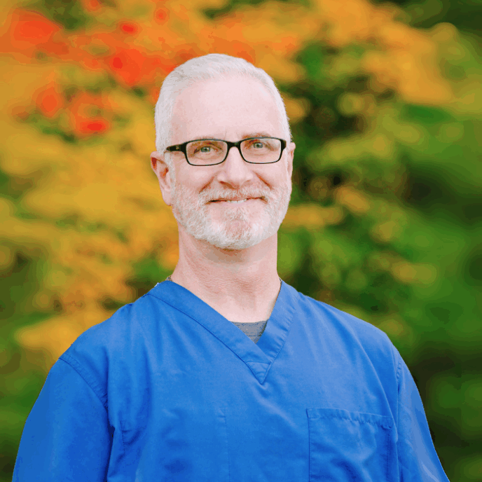 smiling male healthcare professional with white hair and glasses stands outdoors in front of fall foliage. He is wearing blue medical scrubs.