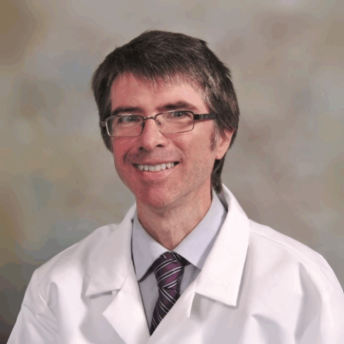 Smiling male clinician with short dark hair and glasses, wearing a white medical coat and striped tie, posed against a neutral background.