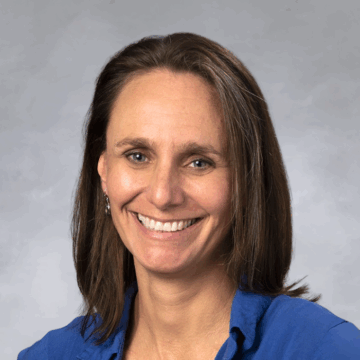 Smiling woman with straight brown hair wearing a bright blue collared shirt and silver earrings, in front of a soft gray studio background.