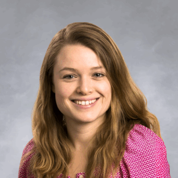 Smiling woman with long, wavy light brown hair wearing a bright pink patterned blouse, photographed against a white background.