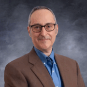 A man with short gray hair and glasses is smiling slightly at the camera. He is wearing a brown blazer over a blue button-up shirt. The background is a neutral cloudy gradient in shades of gray and blue.