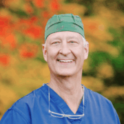 Smiling older man in blue scrubs and a green scrub cap with reading glasses resting on his chest, standing in front of red and yellow foliage.
