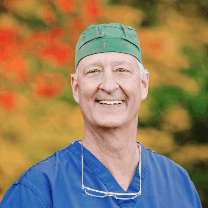 Smiling older man in blue scrubs and a green scrub cap with reading glasses resting on his chest, standing in front of red and yellow foliage.