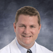 Smiling man with short light brown hair, wearing a white medical coat, light blue shirt, and striped gray tie, posed against a plain white background.