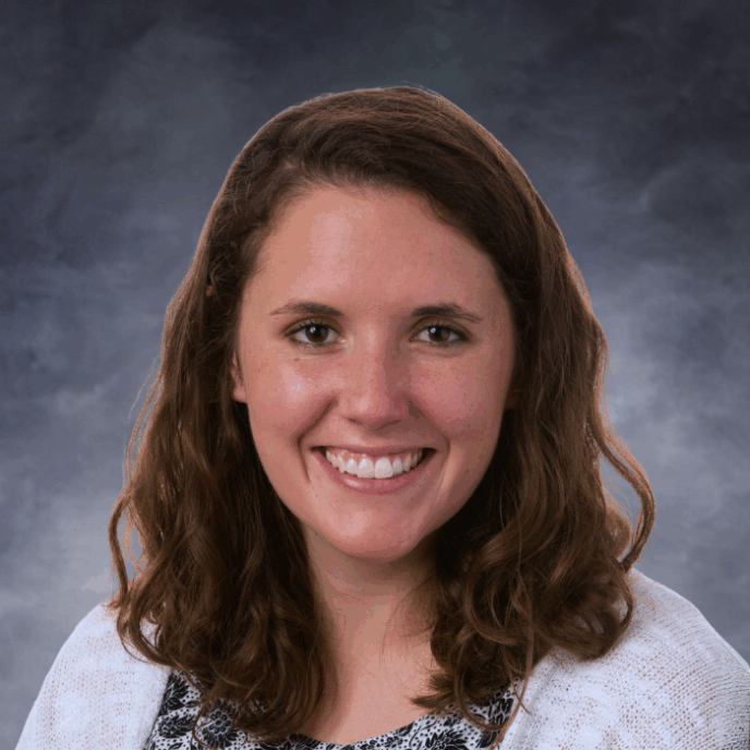 Smiling woman with shoulder-length wavy brown hair wearing a white cardigan over a black and white floral-patterned top, in front of a soft gray studio background.