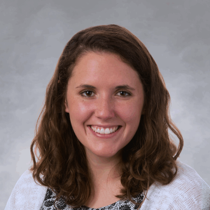 Smiling woman with shoulder-length wavy brown hair wearing a white cardigan over a black and white floral-patterned top, in front of a soft gray studio background.