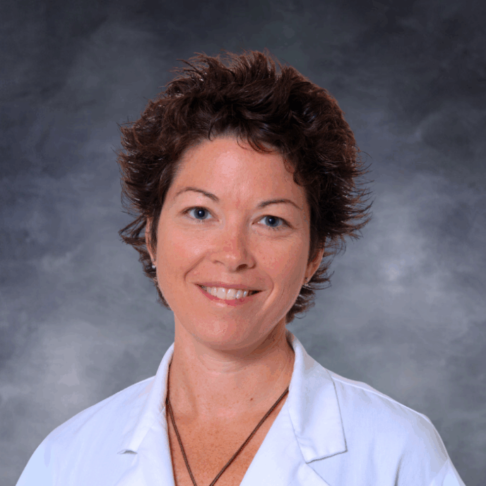 Smiling woman with short, spiky dark brown hair wearing a white medical coat, a turquoise top, and a necklace with a round pendant, photographed against a white background.