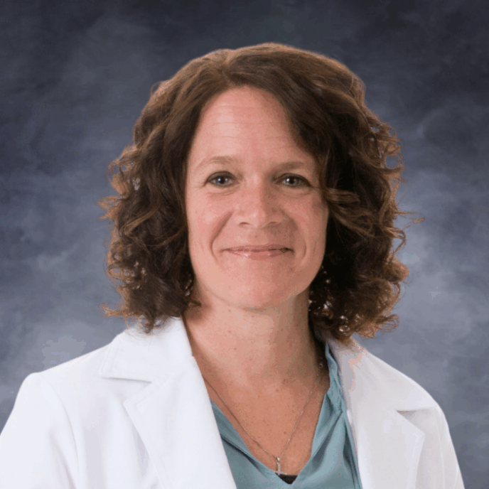 Smiling woman with shoulder-length curly brown hair wearing a white medical coat, a light blue blouse, and a cross necklace, photographed against a white background.