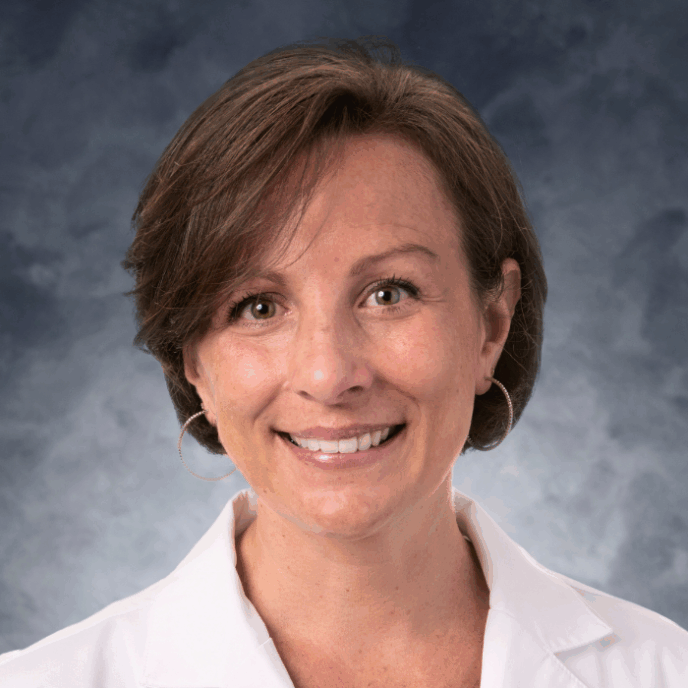 Smiling woman with short brown hair wearing hoop earrings and a white medical coat, in front of a soft gray studio background.