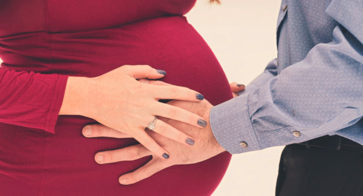 Hands of an expectant couple gently resting on a pregnant belly, with the woman wearing a red dress and the man in a blue button-down shirt