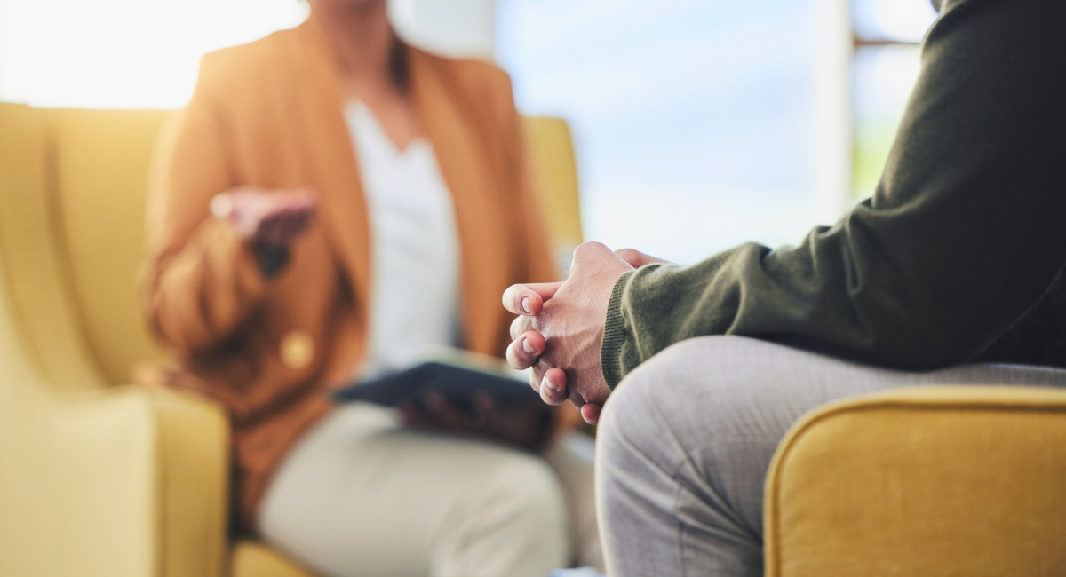 A person sits with hands clasped, speaking with a mental health professional during a counseling session. Both are seated in comfortable chairs in a bright, private office setting.