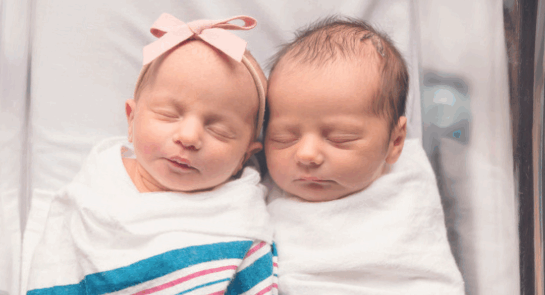 Two newborn twins wrapped in white blankets, sleeping side by side in a hospital bassinet; one wears a pink bow headband