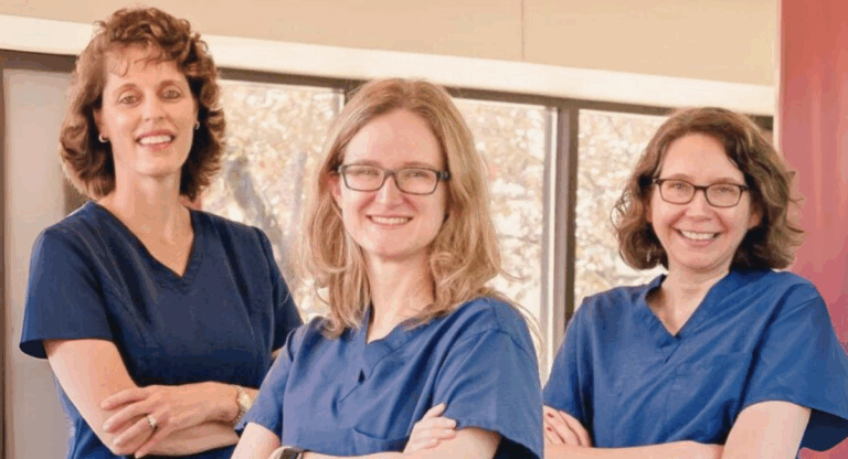 Three OB/GYN providers in navy scrubs standing with arms crossed and smiling in a sunlit room