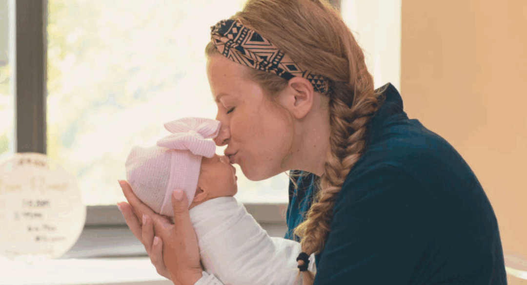 A mother with braided hair and a patterned headband gently kisses her newborn baby, who is wearing a pink bow hat