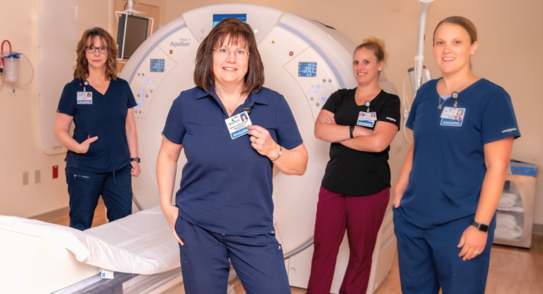 A group of four radiology staff members stands confidently in front of an advanced imaging machine, wearing uniforms and name tags, highlighting their professionalism and teamwork