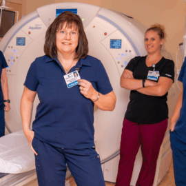 A group of four radiology staff members stands confidently in front of an advanced imaging machine, wearing uniforms and name tags, highlighting their professionalism and teamwork