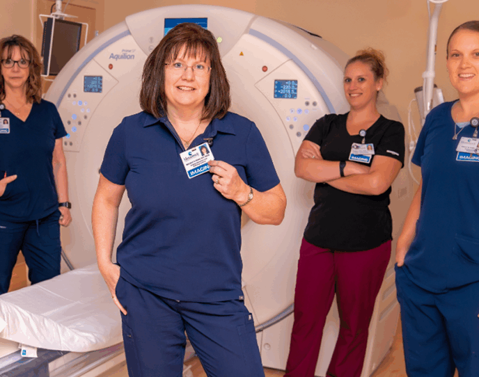 A group of four radiology staff members stands confidently in front of an advanced imaging machine, wearing uniforms and name tags, highlighting their professionalism and teamwork