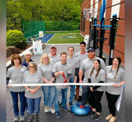 A group of eleven people, all wearing gray shirts, stand outdoors in front of a sports rehabilitation facility. They are smiling, with one person in the center holding a pair of scissors as if preparing to cut a ceremonial ribbon. The background shows a sports court and exercise equipment, emphasizing the focus on sports rehabilitation. The scene is bright and welcoming, indicating a celebratory event, likely the opening or launch of the facility