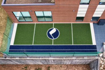 Aerial view of an outdoor sports rehabilitation area with a green and blue synthetic turf, featuring a circular logo in the center. The space is adjacent to a brick building with large windows