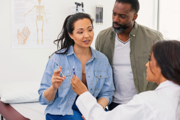 A female patient discussing an injection with her physician while her partner looks on attentively in a clinical setting with a medical chart in the background