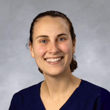 Smiling woman with dark brown hair pulled back, wearing navy blue scrubs, in front of a soft gray studio background.