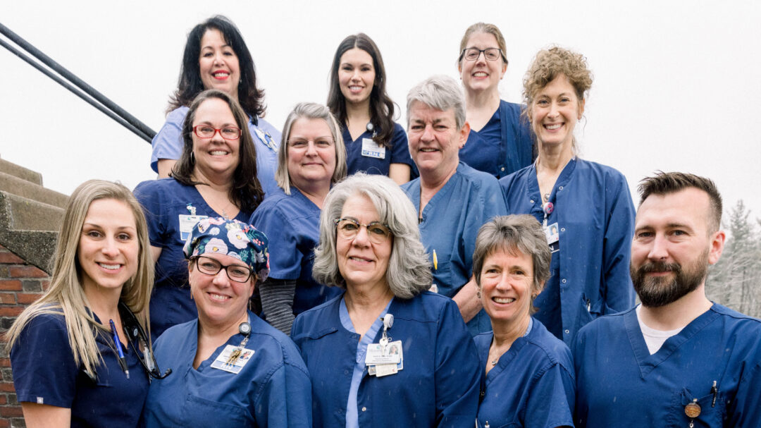 Group of Monadnock Community Hospital nurses standing together outdoors, smiling at the camera. They are wearing navy blue scrubs and ID badges, representing a diverse and dedicated nursing team.