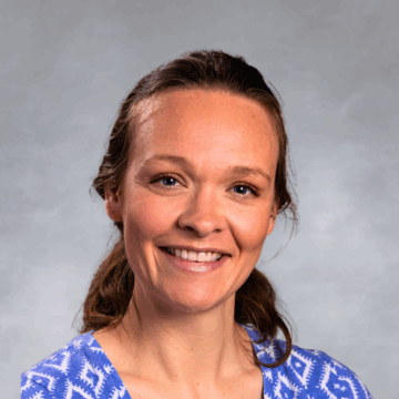 Smiling woman with light brown hair pulled back, wearing a blue and white patterned top, in front of a soft gray studio background.