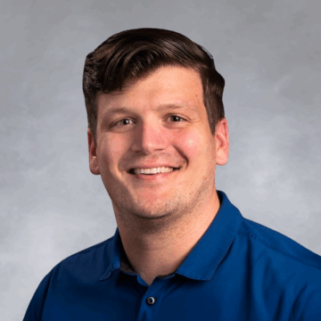Smiling man with short, wavy brown hair wearing a royal blue collared shirt, in front of a soft gray studio background.