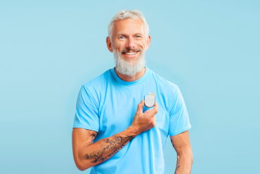 A smiling older man with a beard holding a pacemaker device against his chest, wearing a blue shirt against a plain blue background