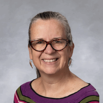 Smiling woman with gray hair pulled back, wearing tortoiseshell glasses, gold earrings, and a striped purple, green, and black sweater, in front of a soft gray studio background.