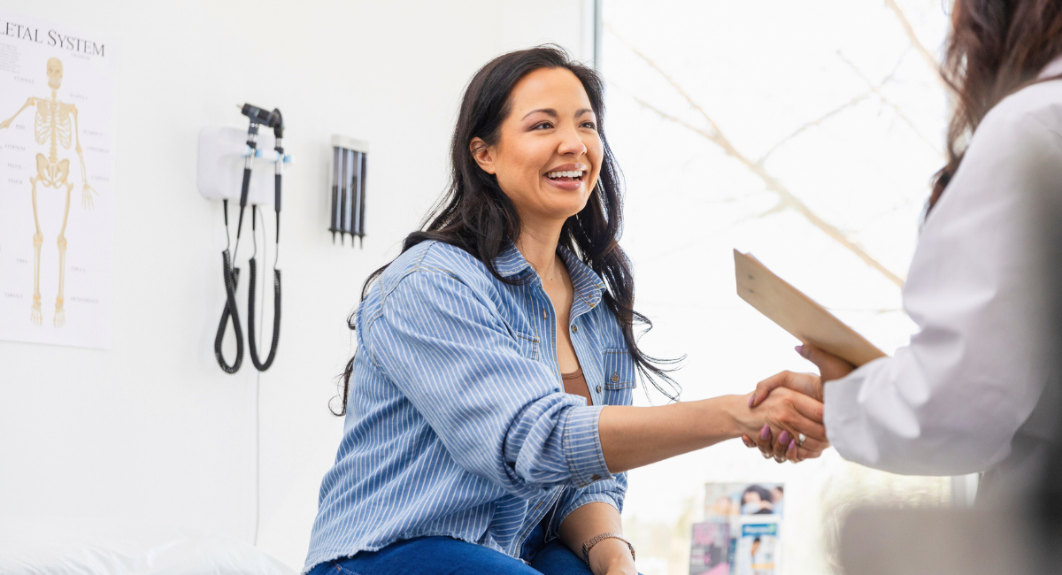Smiling patient shaking hands with a healthcare provider in a bright exam room, with medical tools and a skeletal system chart visible on the wall.