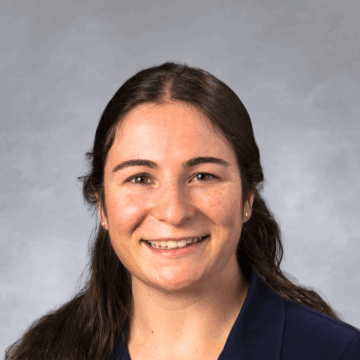 Smiling woman with long, dark wavy hair pulled back, wearing a navy blue collared shirt, in front of a soft gray studio background.