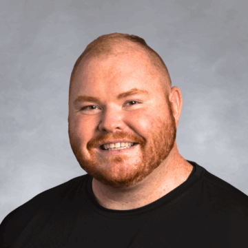 Smiling man with short red hair and beard, wearing a black crewneck shirt, in front of a soft gray studio background.