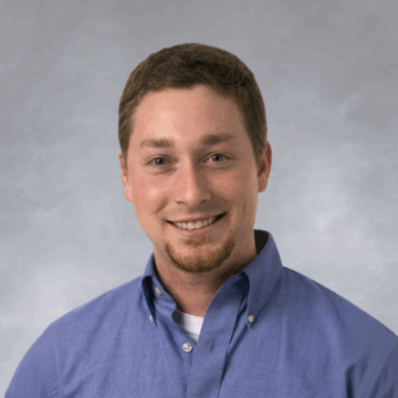 Smiling man with short brown hair and a trimmed goatee, wearing a blue button-down shirt over a white undershirt, photographed against a white background.
