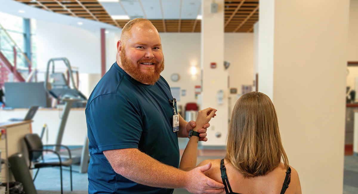 Physical therapist guiding a patient through a shoulder exercise in a rehabilitation space with gym equipment in the background