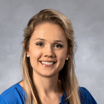 Smiling woman with long blonde hair pulled back, wearing a royal blue top and pearl earrings, in front of a soft gray studio background.