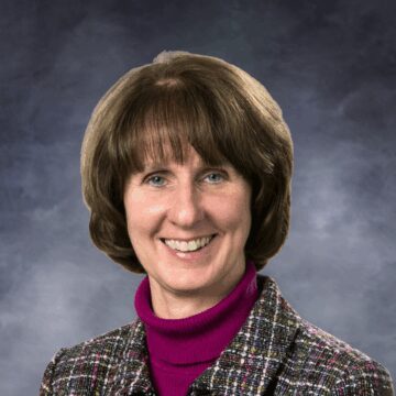 Smiling woman with short brown hair wearing a magenta turtleneck and a multicolored tweed jacket, photographed against a white background.