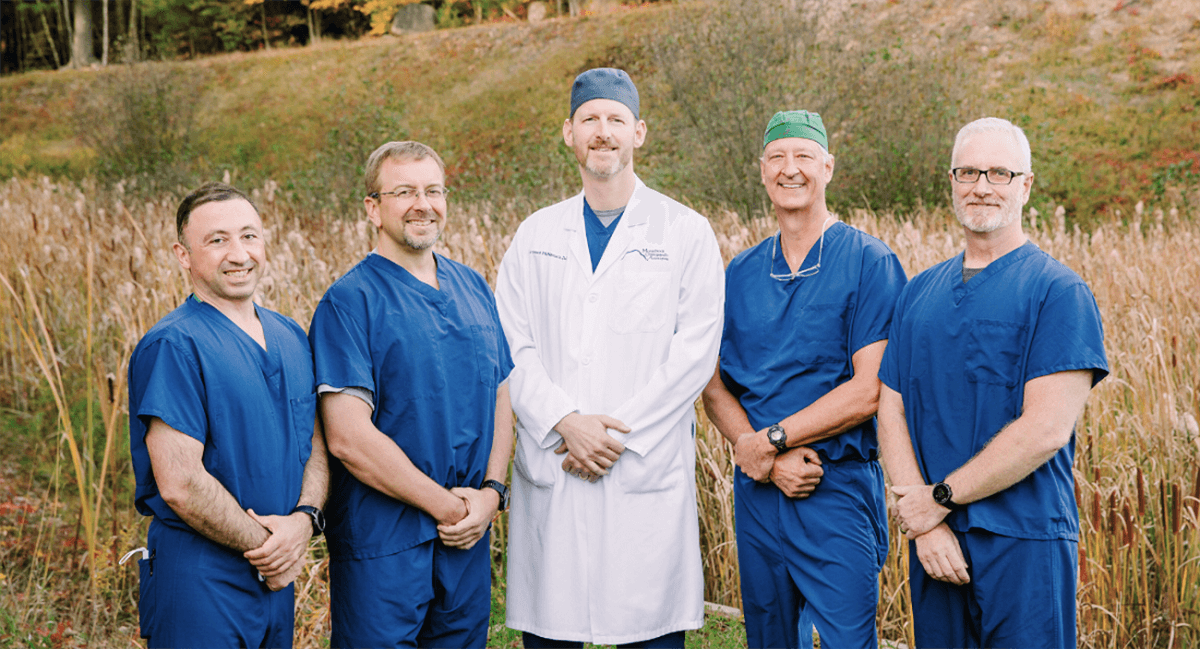 Five orthopedic providers from Monadnock Community Hospital pose outdoors in front of tall grass and autumn trees. Four are wearing blue surgical scrubs, and one is in a white lab coat, all smiling and standing side by side.