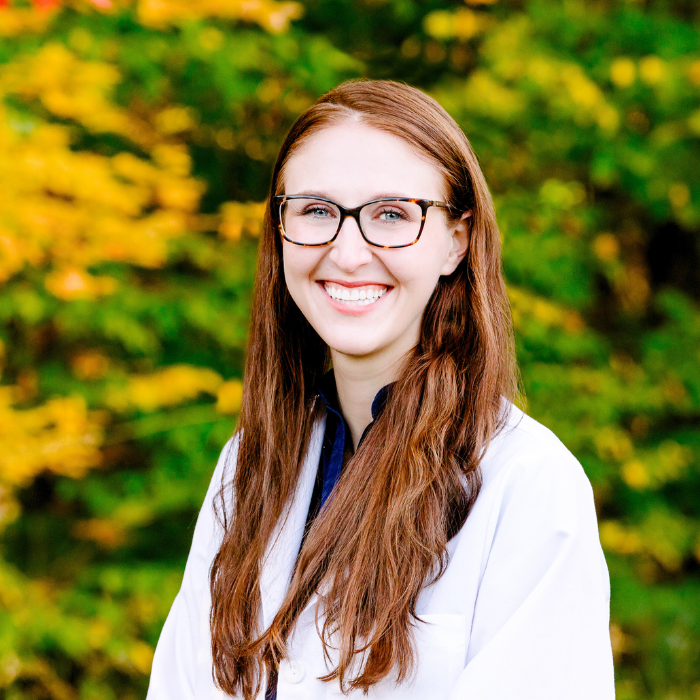 Smiling woman with long auburn hair and glasses, wearing a white coat, standing in front of green and yellow foliage.