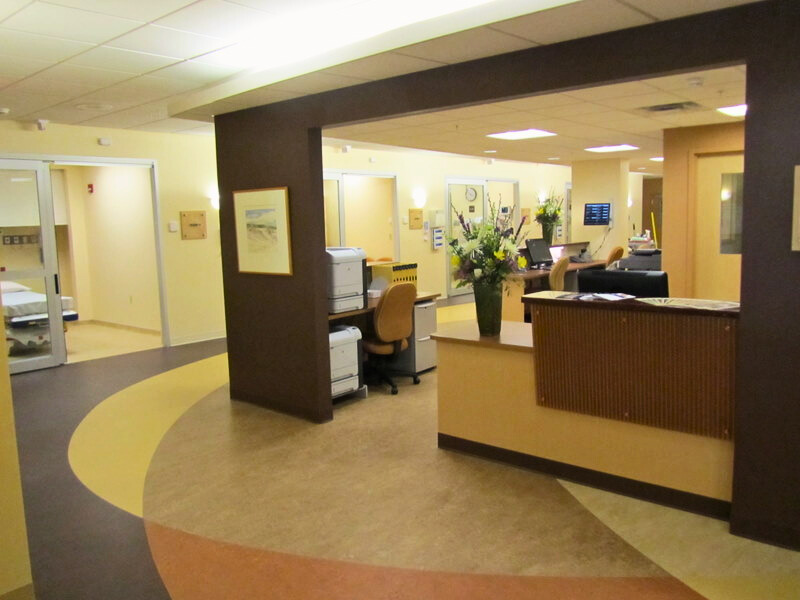 The reception and nurse's station area in the emergency department of Monadnock Community Hospital, showcasing a well-organized workspace with office equipment, floral arrangements, and a view of patient rooms in the background.