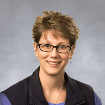 Smiling woman with short, spiky light brown hair and glasses, wearing a black vest over a purple shirt, photographed against a white background.