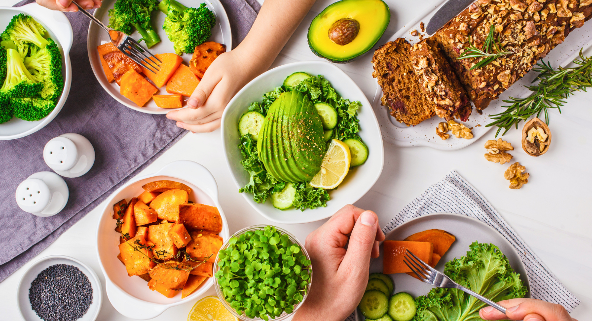 Top-down view of a healthy meal spread with bowls of roasted sweet potatoes, broccoli, leafy green salad with avocado and lemon, a small bowl of microgreens, a plate of nut bread, and hands reaching in to serve, emphasizing balanced, nutritious eating.