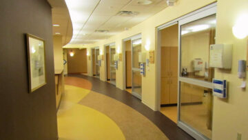 A modern and clean hallway in the emergency department of Monadnock Community Hospital, featuring several private patient rooms with glass doors and well-lit, curved pathways. Alt text for second photo: The reception and nurse's station area in the emergency department of Monadnock Community Hospital, showcasing a well-organized workspace with office equipment, floral arrangements, and a view of patient rooms in the background.