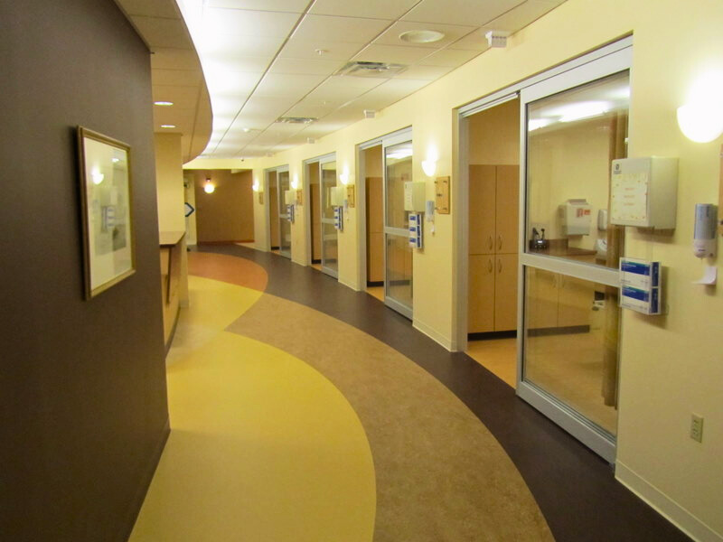 A modern and clean hallway in the emergency department of Monadnock Community Hospital, featuring several private patient rooms with glass doors and well-lit, curved pathways. Alt text for second photo: The reception and nurse's station area in the emergency department of Monadnock Community Hospital, showcasing a well-organized workspace with office equipment, floral arrangements, and a view of patient rooms in the background. 